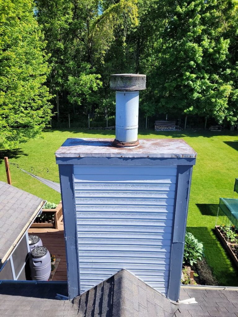 Aerial view of a blue-sided building's chimney with a rusted chase cover, surrounded by trees and lawn.