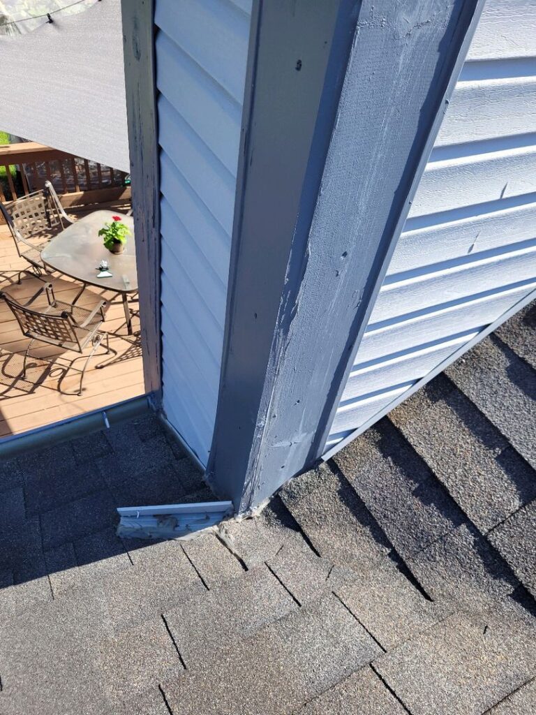 A close-up view of a house corner showing roofing without proper flashing and a detached downspout.