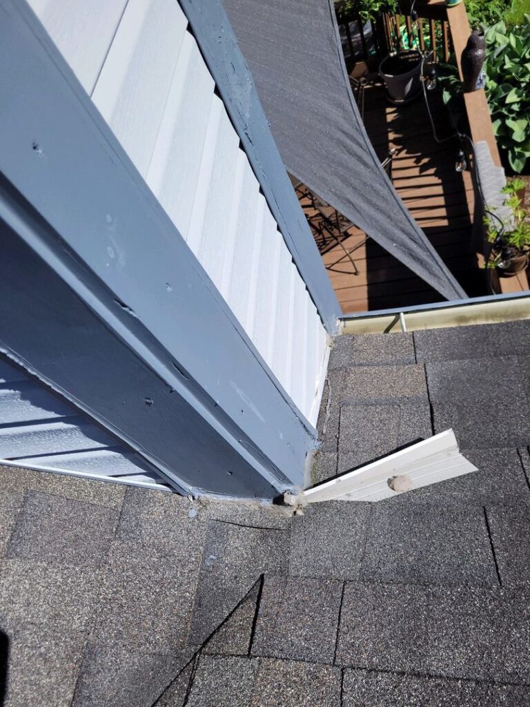 Close-up of a house corner showing a detached gutter downspout, white siding, and a shaded deck area