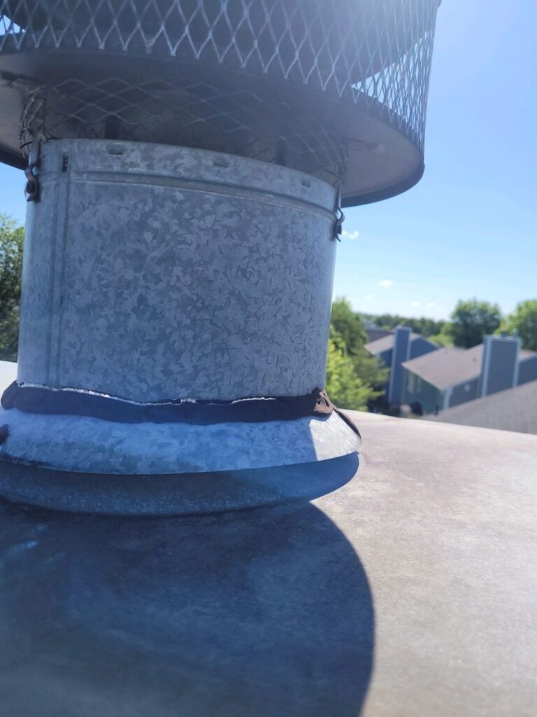 Close-up of a metal chimney cap with mesh on top of a house roof under blue sky.