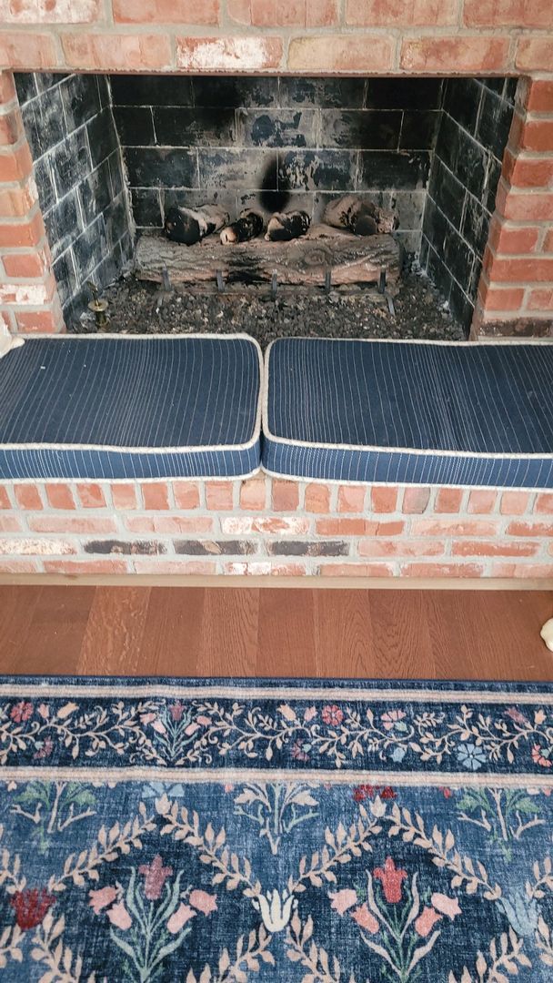 Brick fireplace with soot, two striped cushions in front, and a floral-patterned rug on hardwood floor