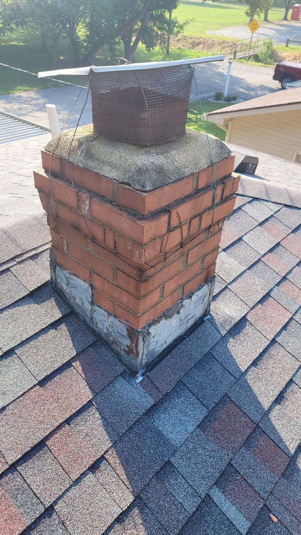 Brick chimney with a mesh cap on top and weathered paint at the base, set against a residential rooftop