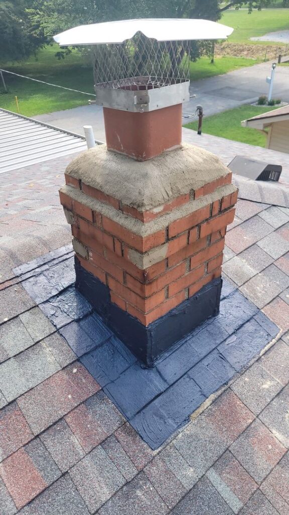 Brick chimney with metal cap and dark sealant on its base on a shingled rooftop