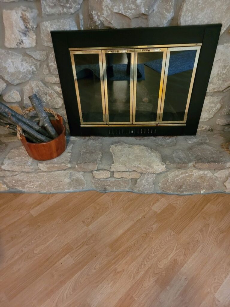 A home's stone wall fireplace with brass-trimmed doors and a bucket of firewood on the side.