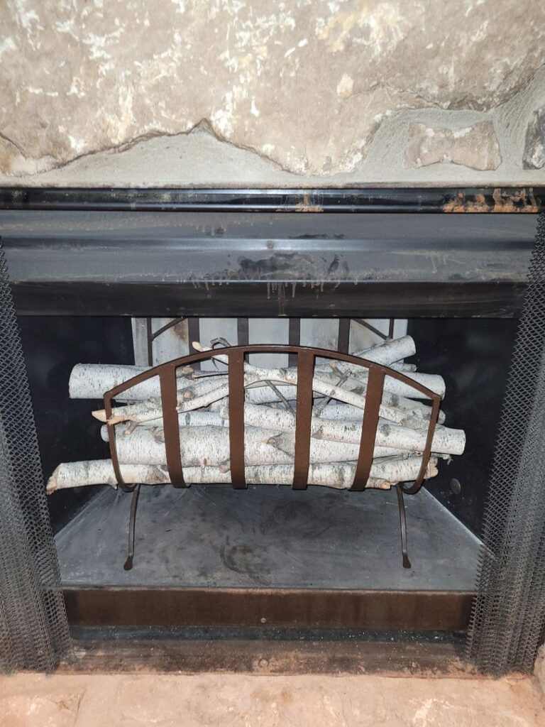 An unlit fireplace with a black frame and mesh screen containing artificial logs arranged on a grate.