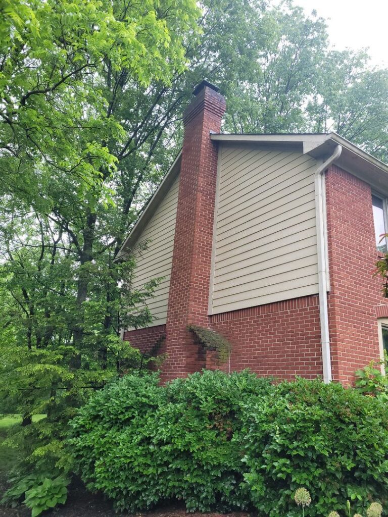 A two-story house with beige siding and a tall brick chimney, set against a backdrop of green trees and shrubs under a cloudy sky.