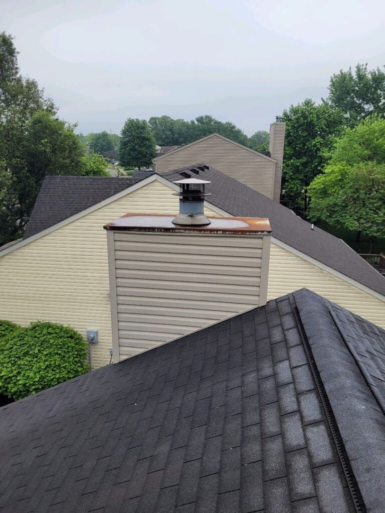 A rusted chimney chase cover on top of a house with asphalt shingle roofing in a suburban neighborhood.