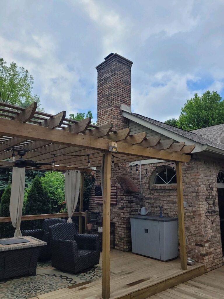 A backyard scene with a tall brick chimney, outdoor furniture under a wooden pergola, and a generator on the deck.