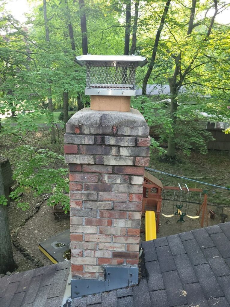 A brick chimney with metal cap on top of a house's shingled roof, surrounded by trees and overlooking a backyard playground.