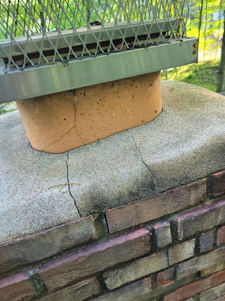 Close-up image of a cracked chimney crown with a metal cap on top of a brick chimney