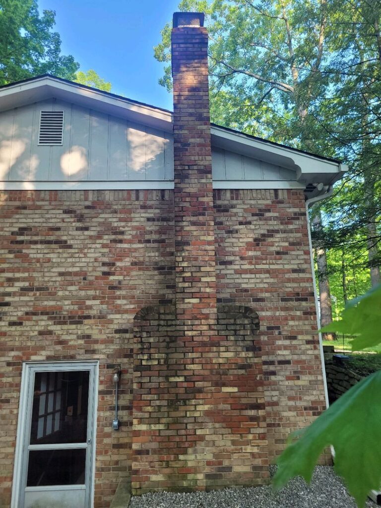 A brick chimney with discoloration on a house with white siding and a window, indicating potential water seepage issues