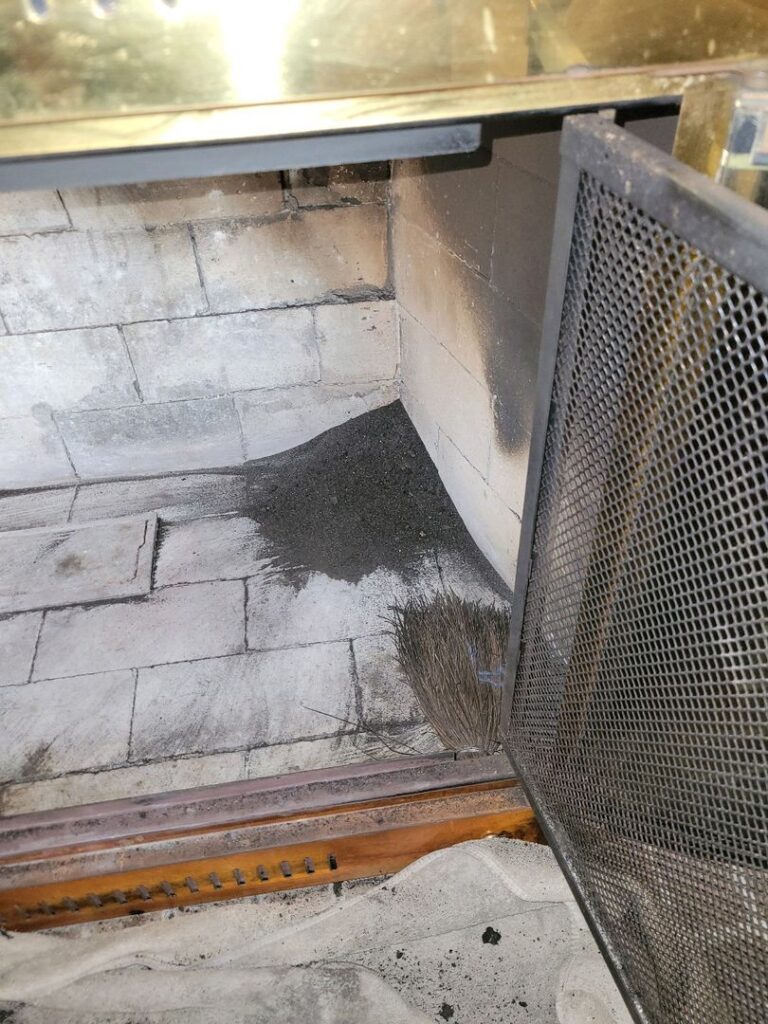 Interior view of a fireplace with ash on the bottom and a broom leaning against the side.