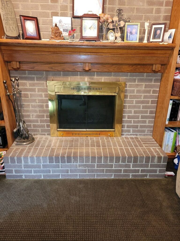 A brick fireplace with a wooden mantel decorated with photos, a clock, and other items, along with fireplace tools on the side.