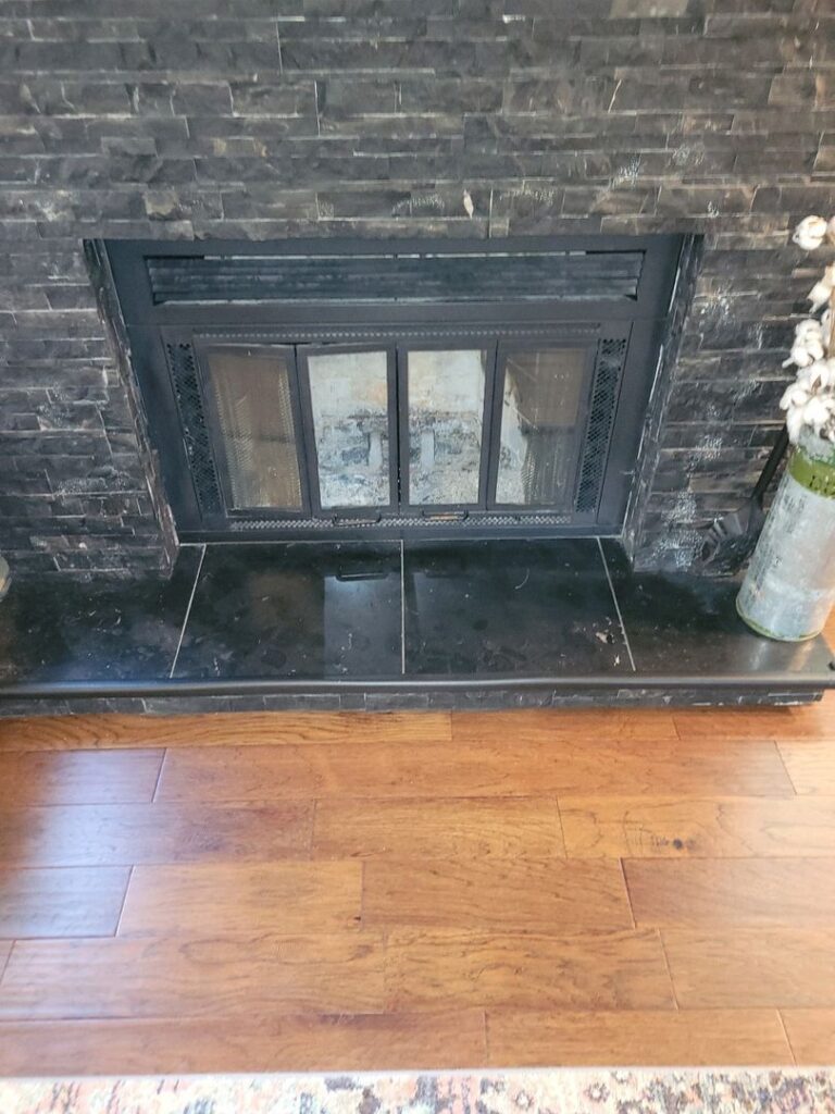 A prefabricated fireplace with glass doors and ash residue inside, set in a black slate surround against a hardwood floor