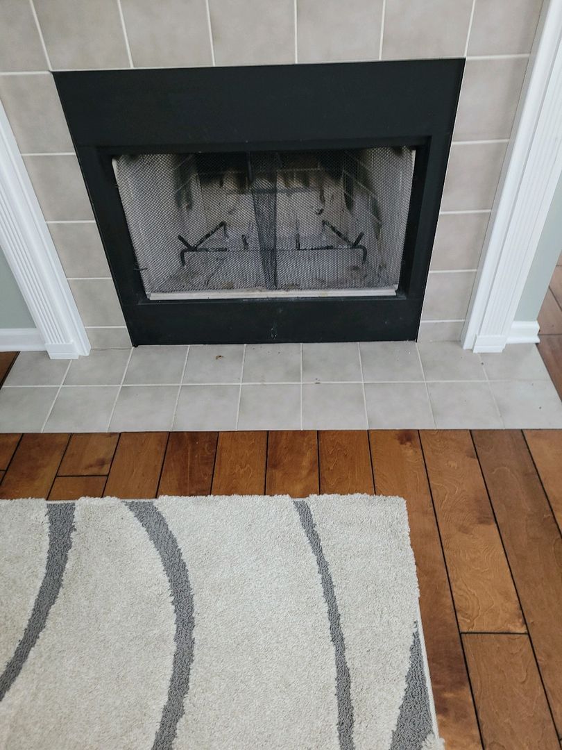 Image of an empty fireplace with a black frame on beige tiles, adjacent to wooden floorboards and a grey tiled area with a patterned rug