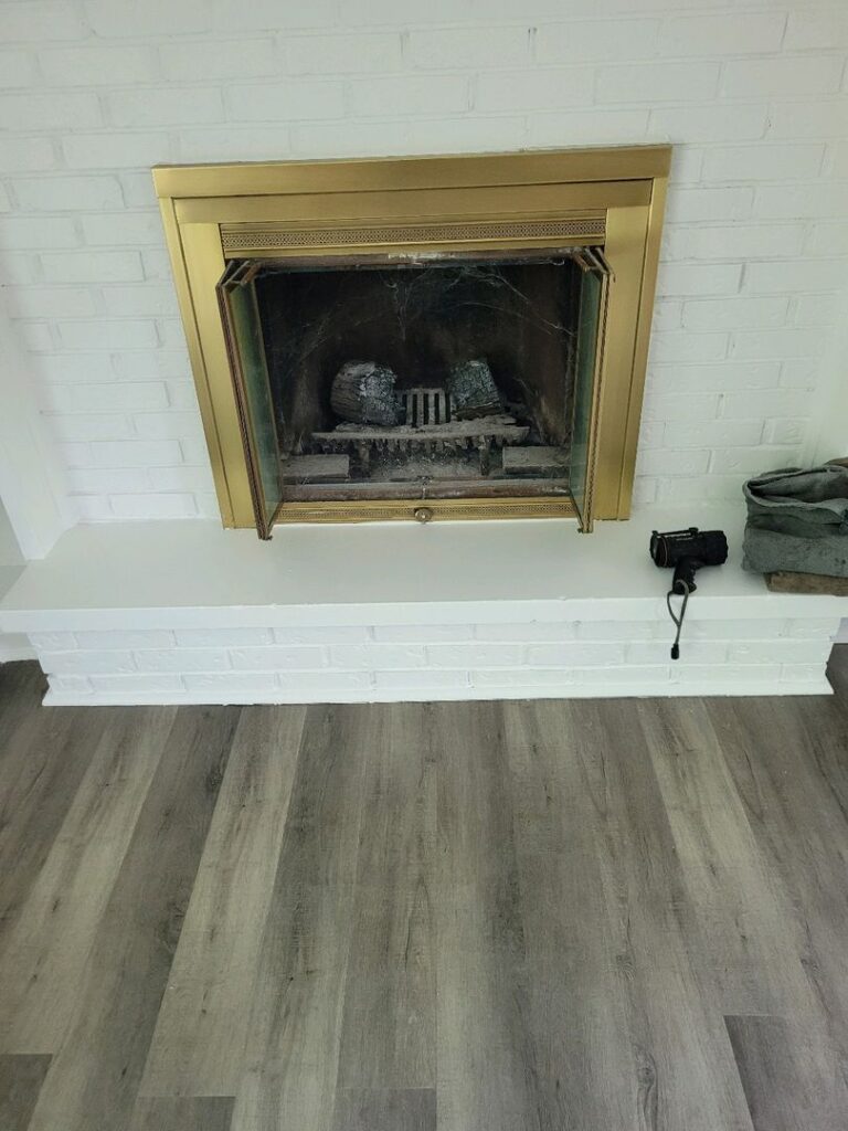 White painted brick fireplace with gold trim and artificial logs inside, next to a black camera on grey wood-patterned flooring.