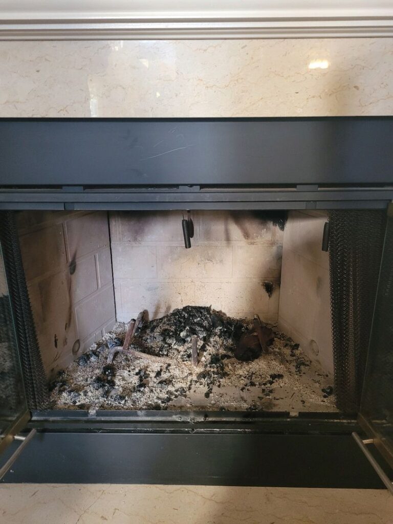 An unlit fireplace filled with ashes and remnants of wood, framed by marble, before cleaning