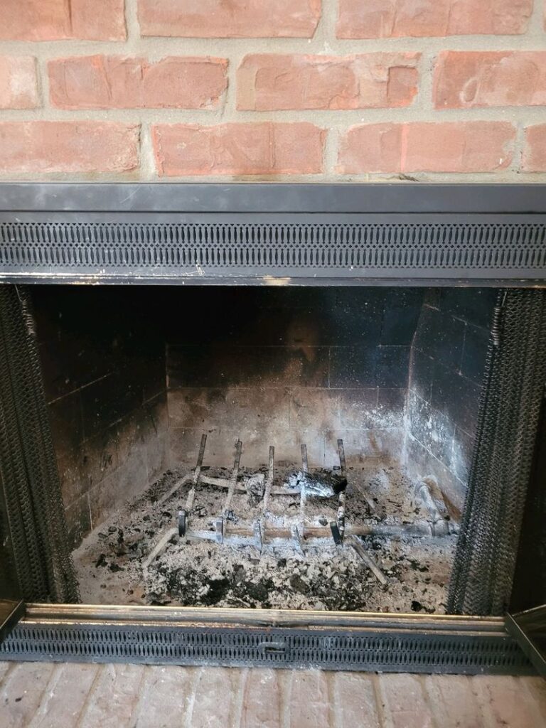 A dirty brick fireplace with a metal grate and accumulated ash, suggesting recent use and need for cleaning.