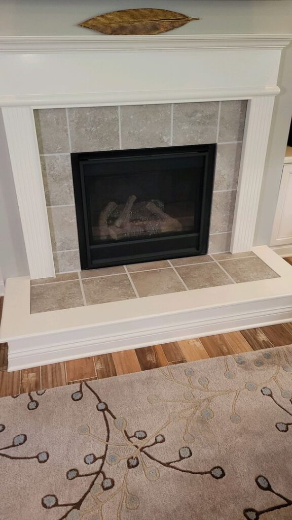 A gas fireplace with stone tiles and a white mantel, decorated with a wicker fish wall art, set against hardwood floors and a patterned beige carpet.