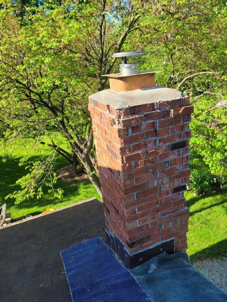 Brick chimney with metal cap on rooftop surrounded by trees and clear blue sky