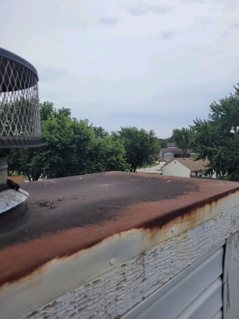 Rusted chase cover on top of a chimney with a metal flue cap in front of trees and houses under an overcast sky.